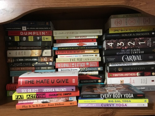 Image shows several stacks of books on a wooden bookshelf, somewhat messy, in no discernible order. Titles include a variety of fiction (The Hate U Give, 4321, John Dies at the End, etc) and non-fiction (Sex Object, Every Body Yoga, Too Fat, Too Slutty, Too Loud, etc).