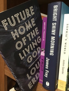 Image shows several books on a shelf, with Future Home of the Living God displayed prominently on the left, sticking out from the other books, as if about to be removed from the shelf. Other books visible include Fruit from Brian Francis and Sophie's World from Jostein Gaarder.