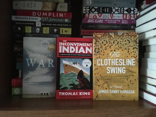 Image displays several stacks of books in the background and three books, covers facing out, in the foreground. Those three books are American War (cover is dominated by a sky full of swirling clouds. In the bottom right corner is a small, white, vacant-looking building), The Inconvenient Indian (cover is red with bold white text and includes an illustration of a Native man wearing a feathered headdress and carrying a spear while looking out over the water at a massive ship approaching), and The Clothesline Swing (a yellow cover with large white text and a multi-colour paisley-esque pattern across the top).