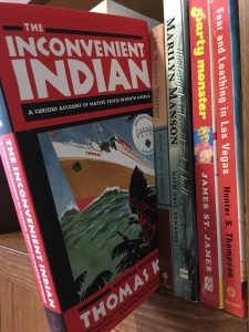Image displays a row of books on a wooden bookshelf. The Inconvenient Indian is pulled out and tilted slightly over the edge.
