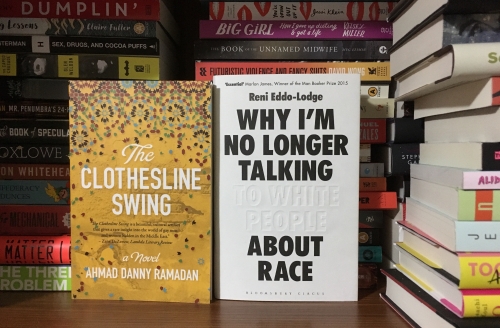 Image displays several stacks of books on a wooden bookshelf. In the foreground are two books with their covers facing out: The Clothesline Swing (a yellow cover with large white text and a multi-colour paisley-esque pattern across the top) and Why I'm No Longer Talking to White People About Race (a white cover with large, bold, black text. The words "to White People" are white and debossed).
